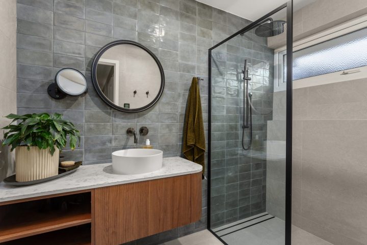 Modern bathroom design featuring a round mirror, green tiled wall, sleek shower enclosure, wooden vanity with a white basin, and a potted plant, reflecting Cornerstone Property Group's renovation style and quality.