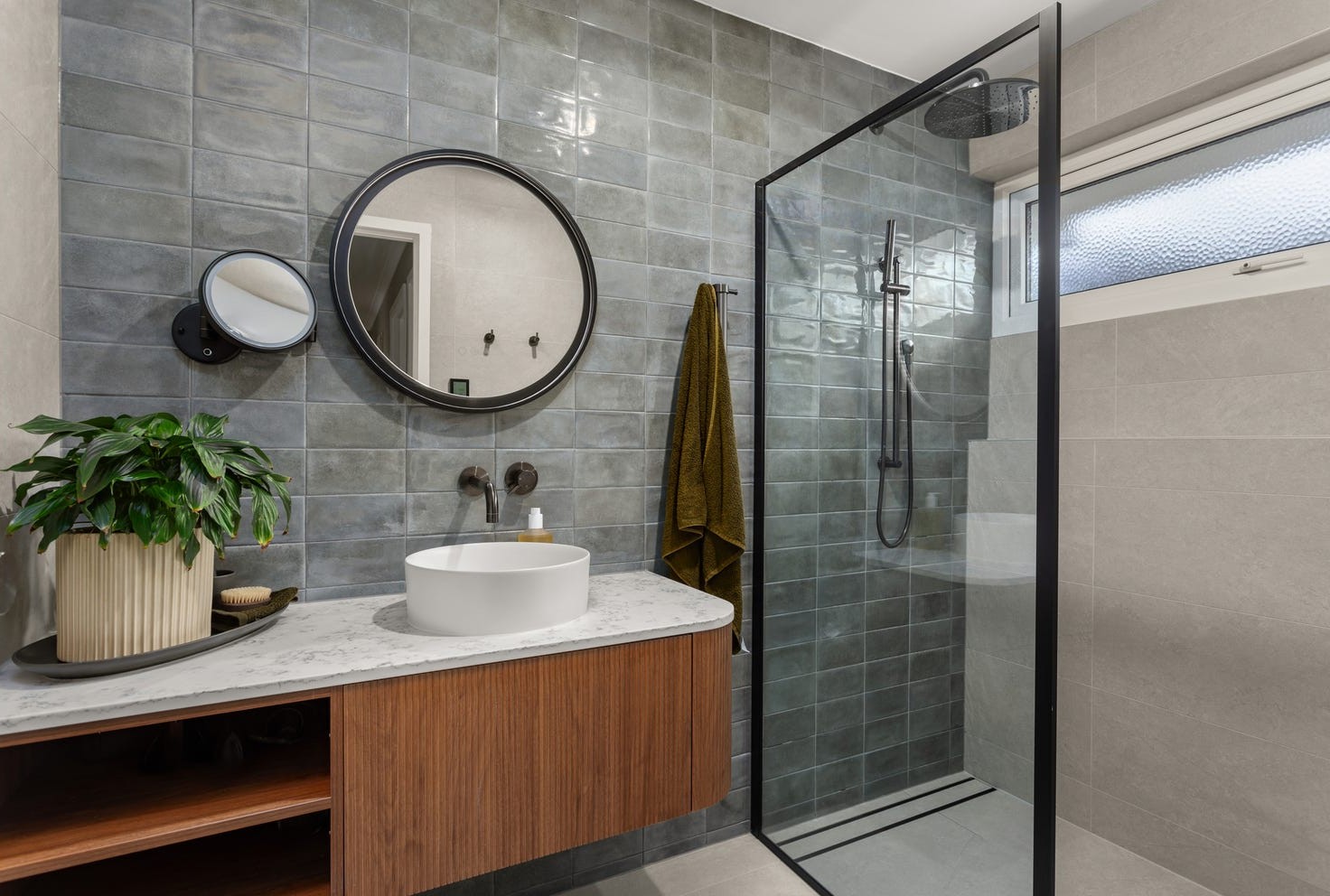 Modern bathroom interior featuring gray tiled walls, round black mirror, white vessel sink, wooden vanity, and glass shower enclosure, highlighting stylish hardware finishes for home renovation.