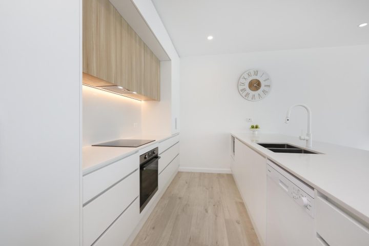 Modern kitchen design featuring sleek white cabinetry, wooden overhead storage, built-in oven, and minimalist clock, showcasing a stylish and functional renovation space by Cornerstone Property Group.