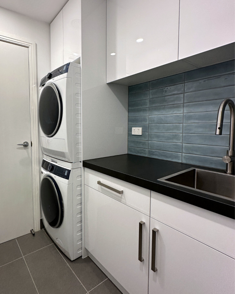 Compact laundry space featuring stacked washer and dryer, sleek cabinetry, black countertop, and stylish blue tiled backsplash.